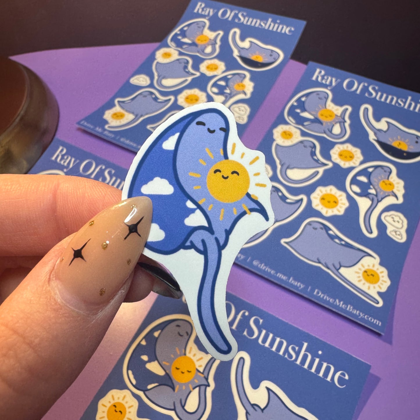 Hand holding a blue shark sticker with a sun and cloud design, surrounded by 'Ray Of Sunshine' branded stickers.