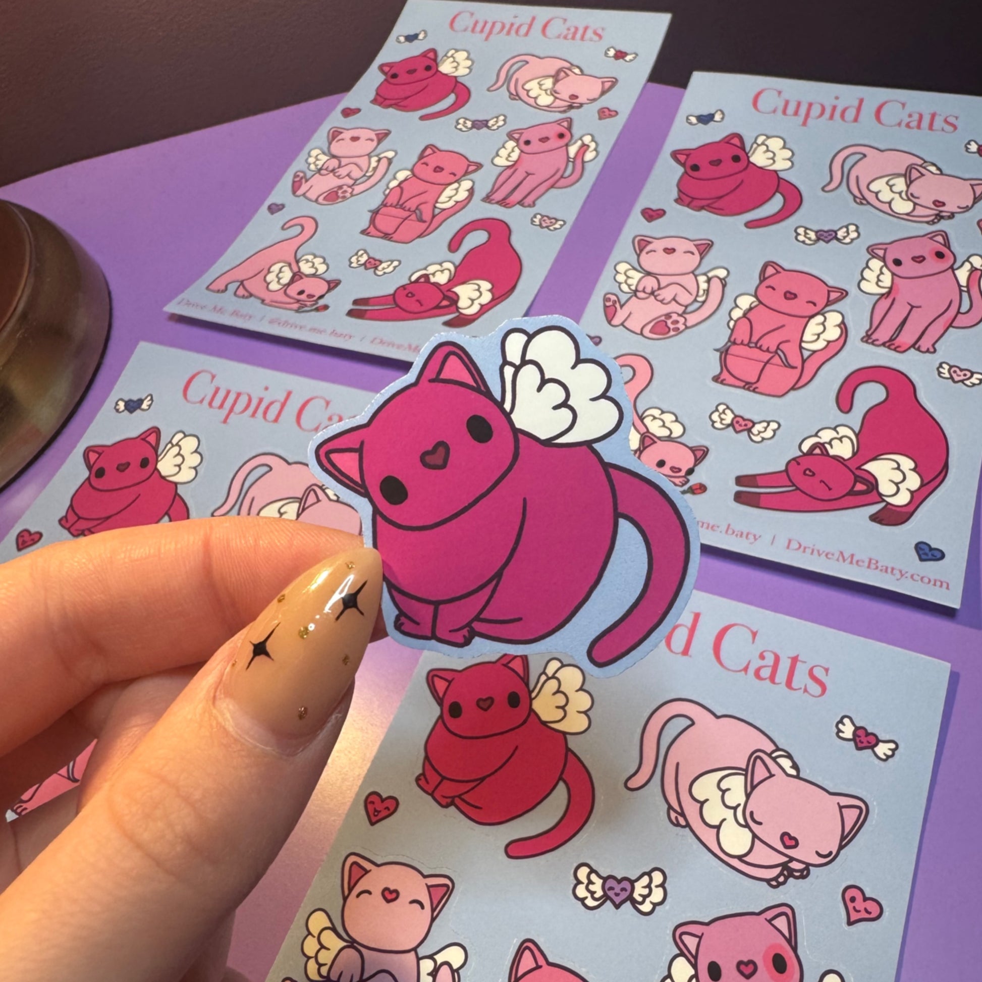 Cupid Cats stickers on a purple surface with a hand holding one sticker.
