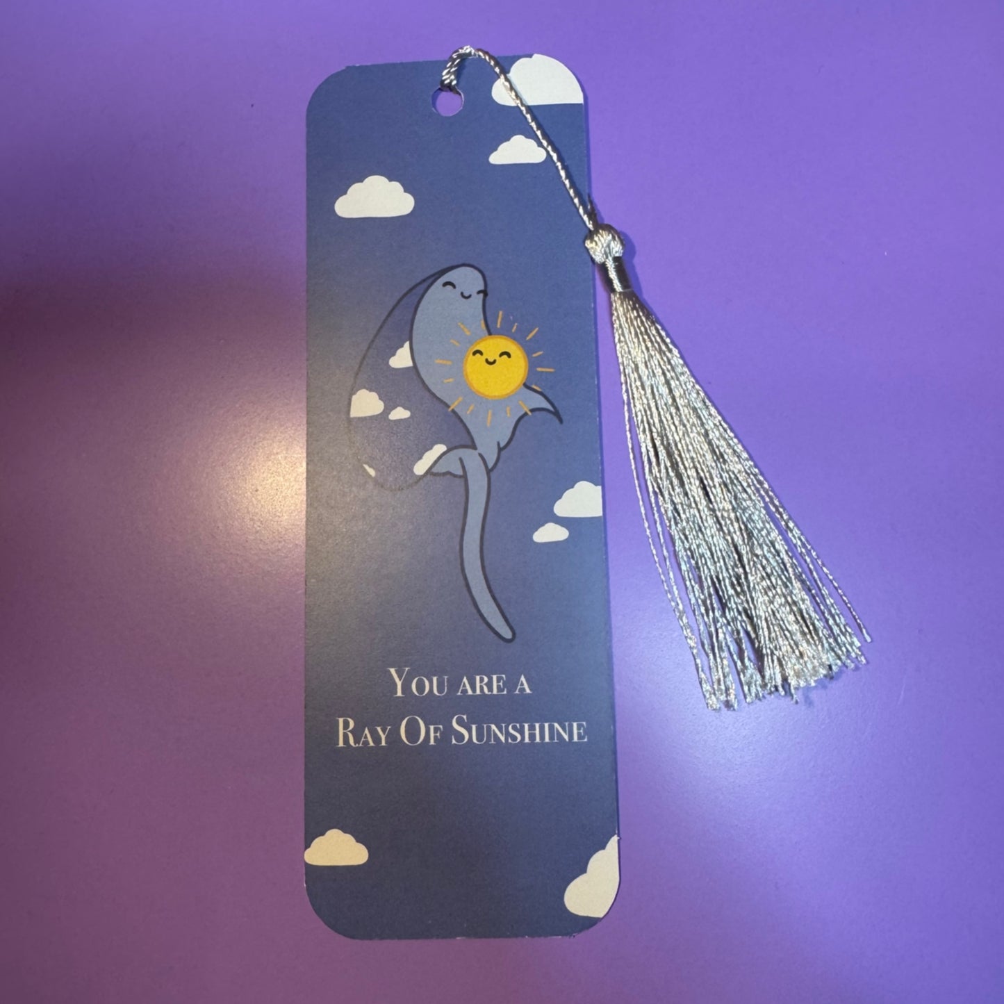 Bookmark with a tassel featuring a cartoon ray and sun design on a purple background.