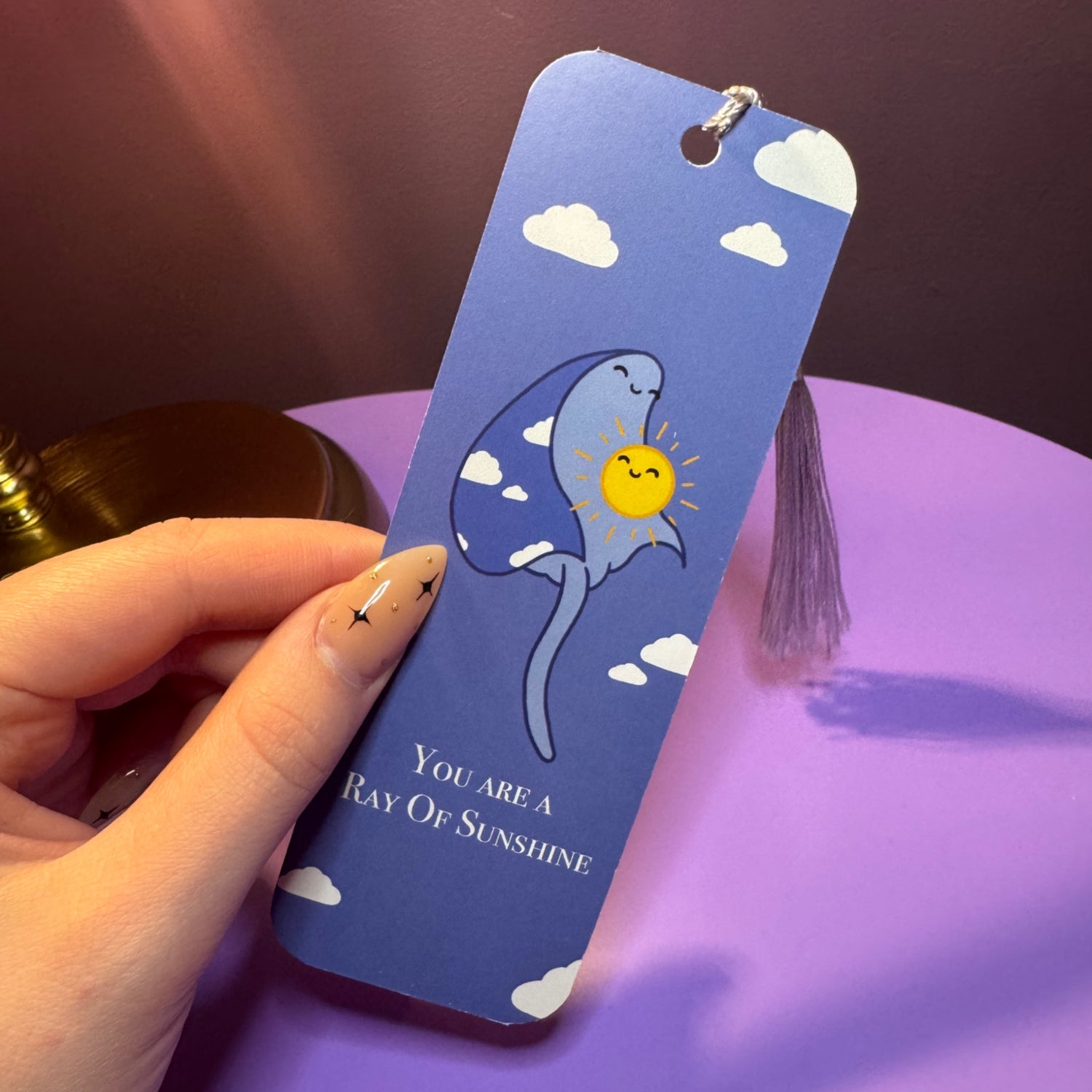 Hand holding a blue bookmark with a ray illustration and text on a purple surface
