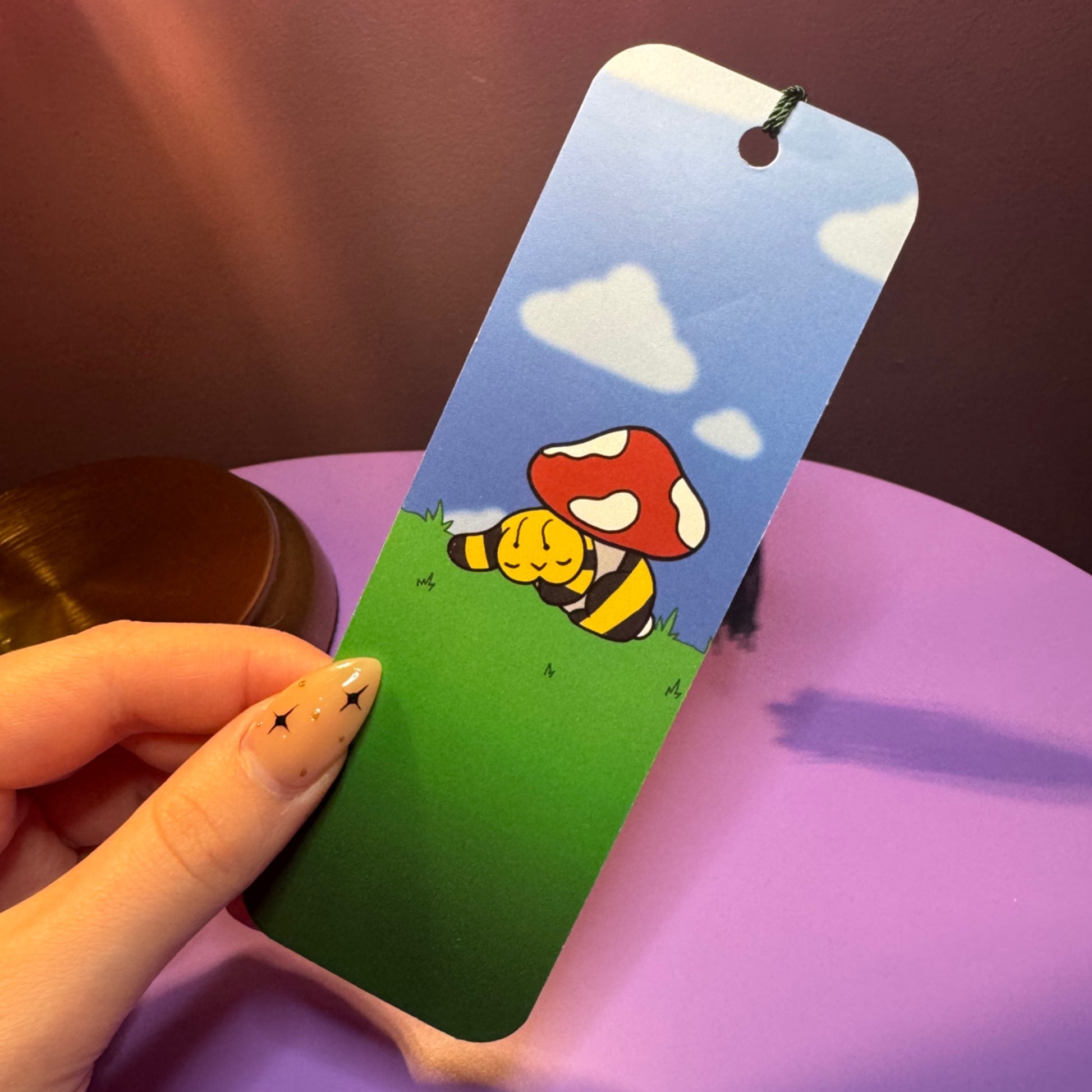 Bookmark with cartoon bee and mushroom design held by a hand on a purple surface.