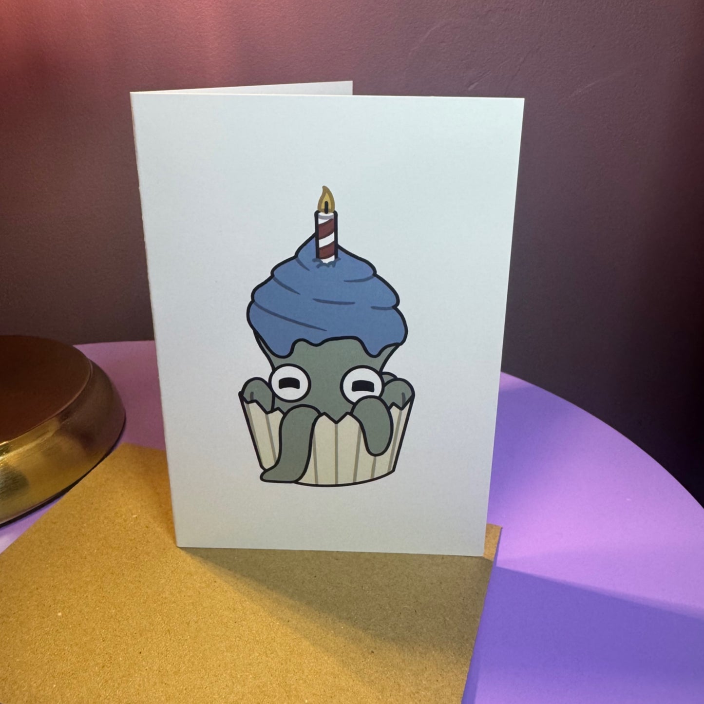 Card with a cartoon octopus disguised as a cupcake featuring blue frosting and a candle on a colourful background.