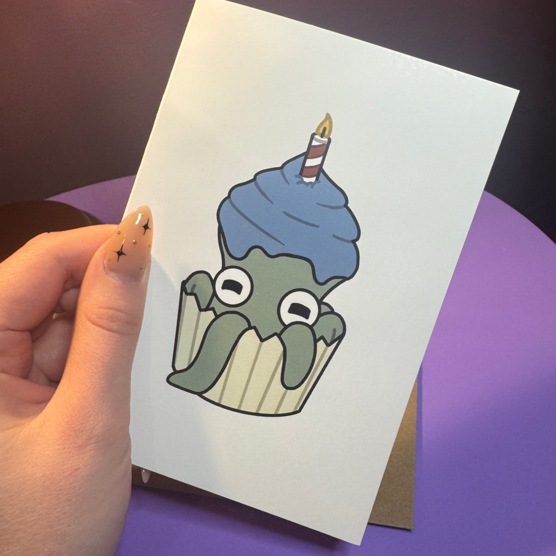 Hand holding a card with a cartoon octopus cupcake design on a purple surface
