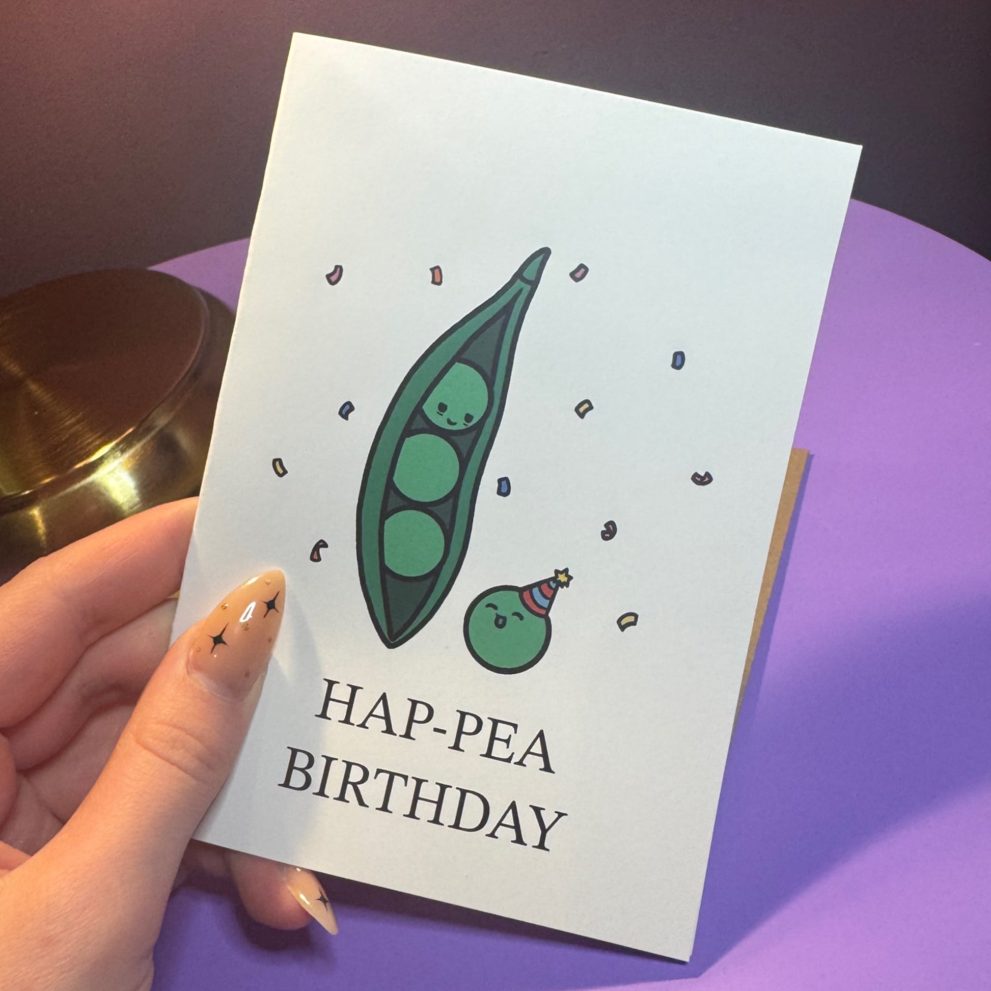 Hand holding a card with a pea pod illustration and 'HAP-PEA BIRTHDAY' text on a purple surface.
