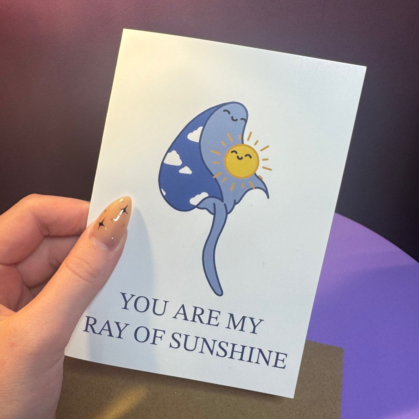 Hand holding a card with a cartoon ray and sun illustration and text 'You are my ray of sunshine'.