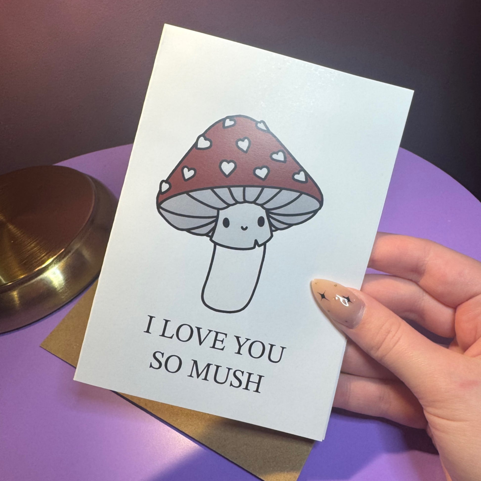 Greeting card with a mushroom illustration and 'I love you so mush' text, held by a hand on a purple surface.